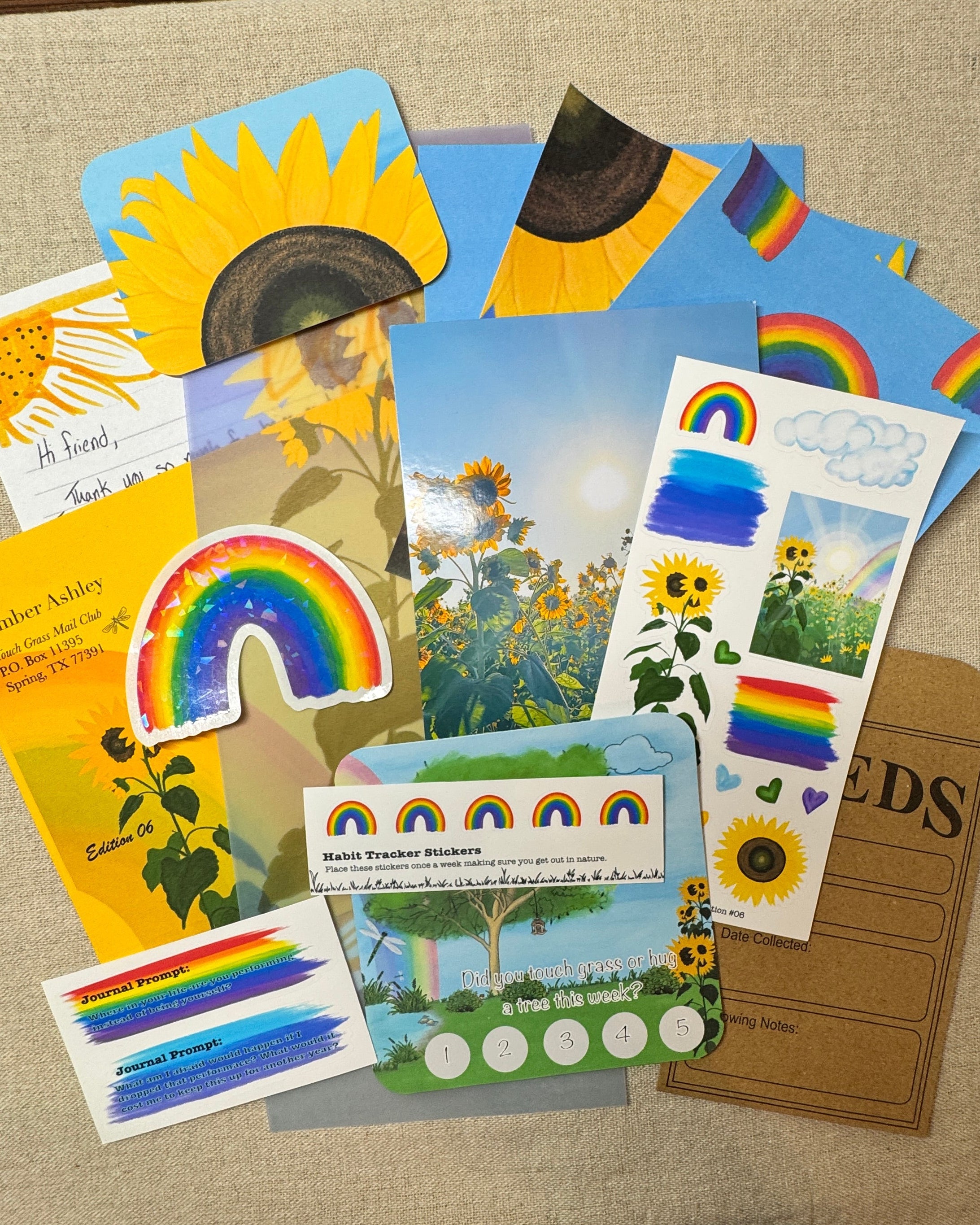 A flat lay of colorful sunflower and rainbow-themed cards, stickers, and a small kraft paper card on a beige fabric surface.