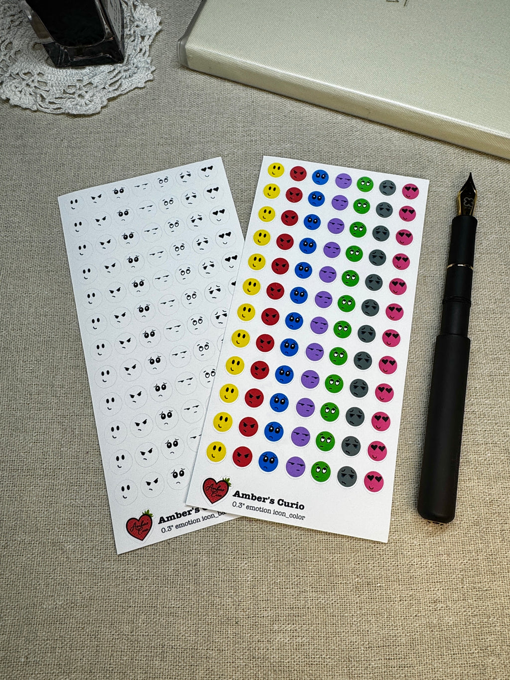 Two sheets of stickers with emotion designs on a linen background next to a pen.