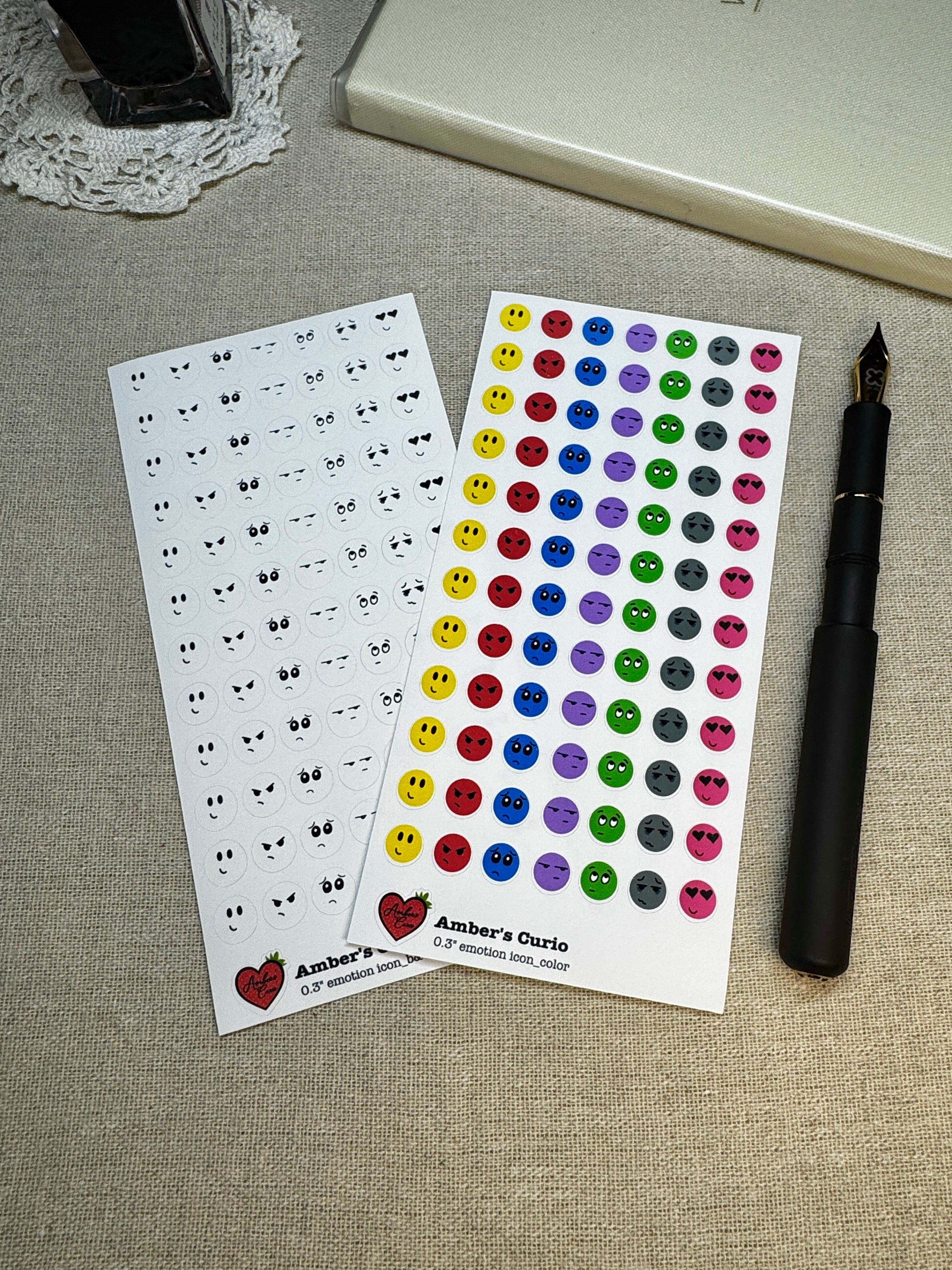 Two sheets of stickers with emotion designs on a linen background next to a pen.