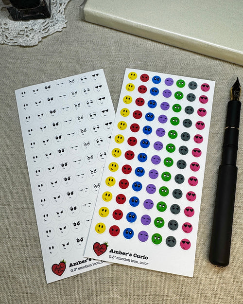 Two sheets of stickers with emotion designs on a linen background next to a pen.