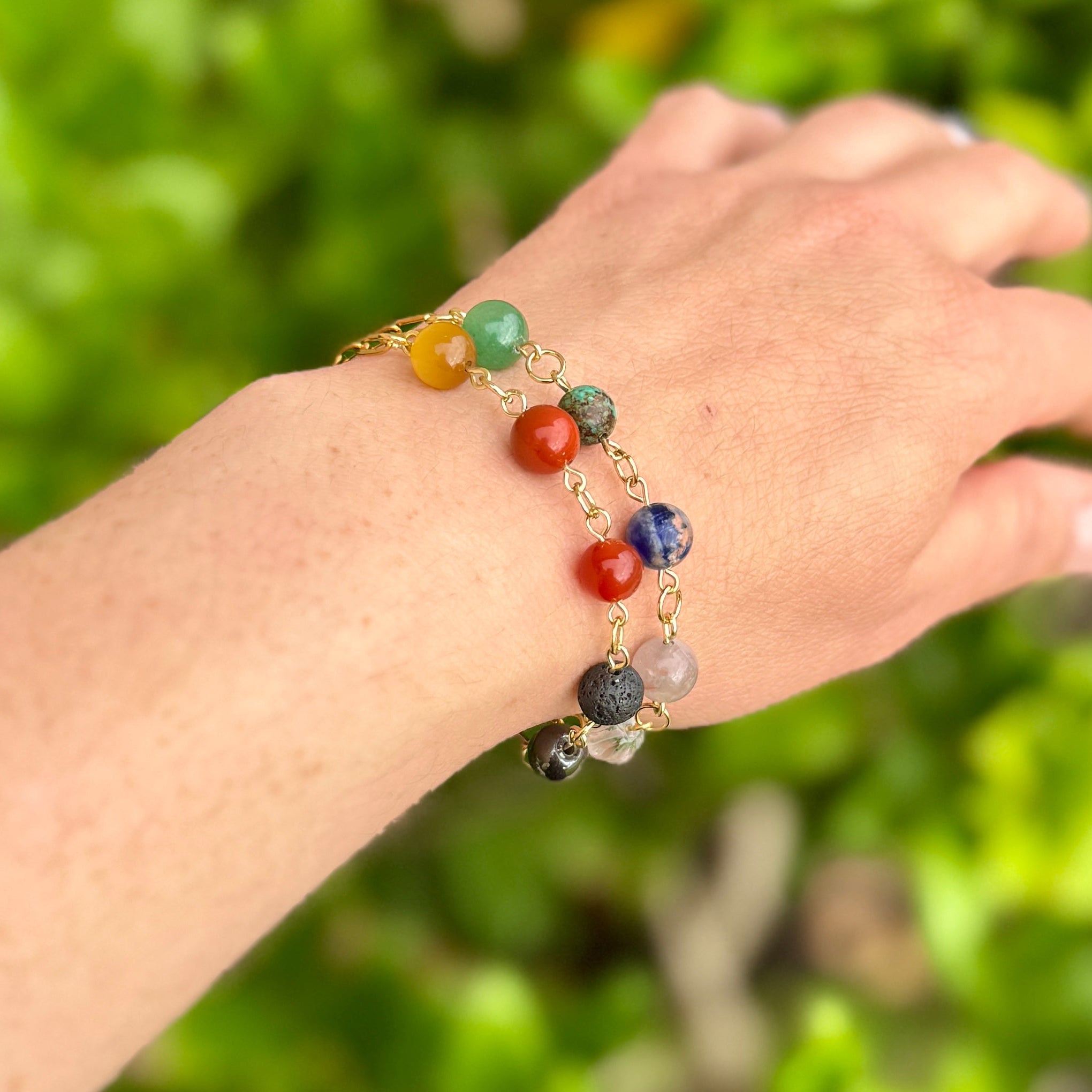 Hand wearing a chakra beaded bracelet in gold against a green leafy background