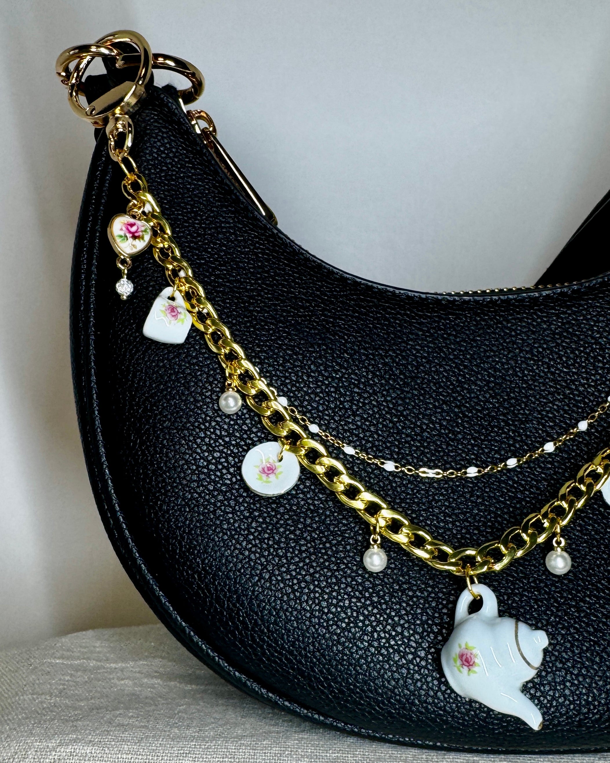 Black handbag with gold chain and ceramic teapot charm on a neutral background
