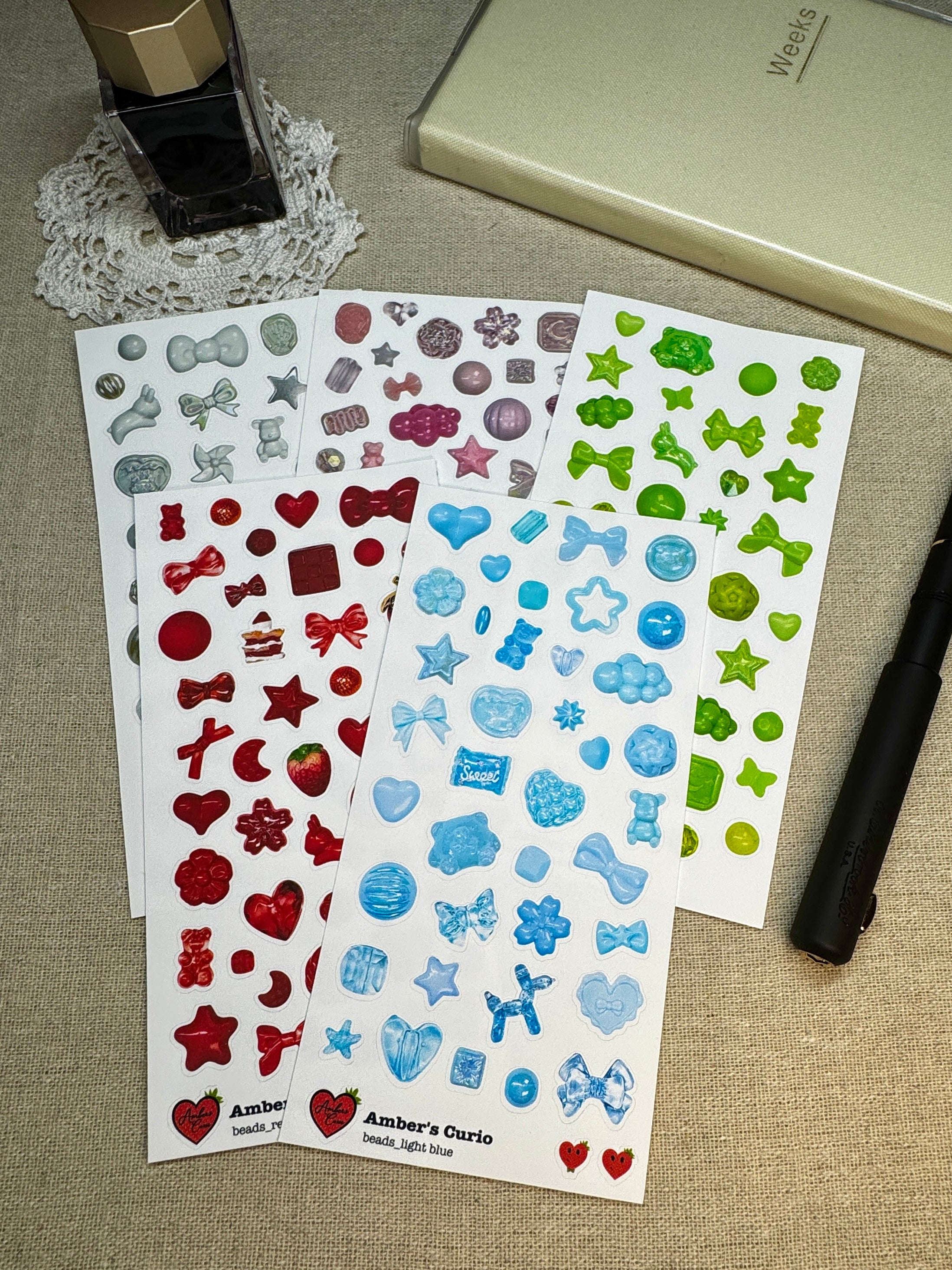 Various colored sticker sheets of photographed beads and charms printed on premium matte paper photographed next to a black fountain pen, journal and fountain ink 
