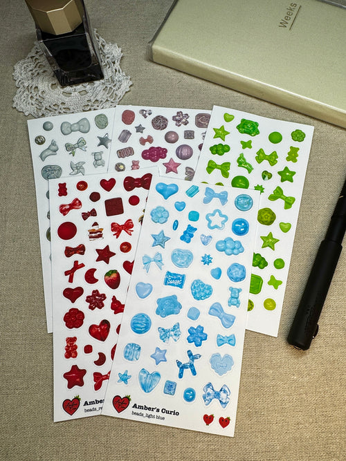Various colored sticker sheets of photographed beads and charms printed on premium matte paper photographed next to a black fountain pen, journal and fountain ink 