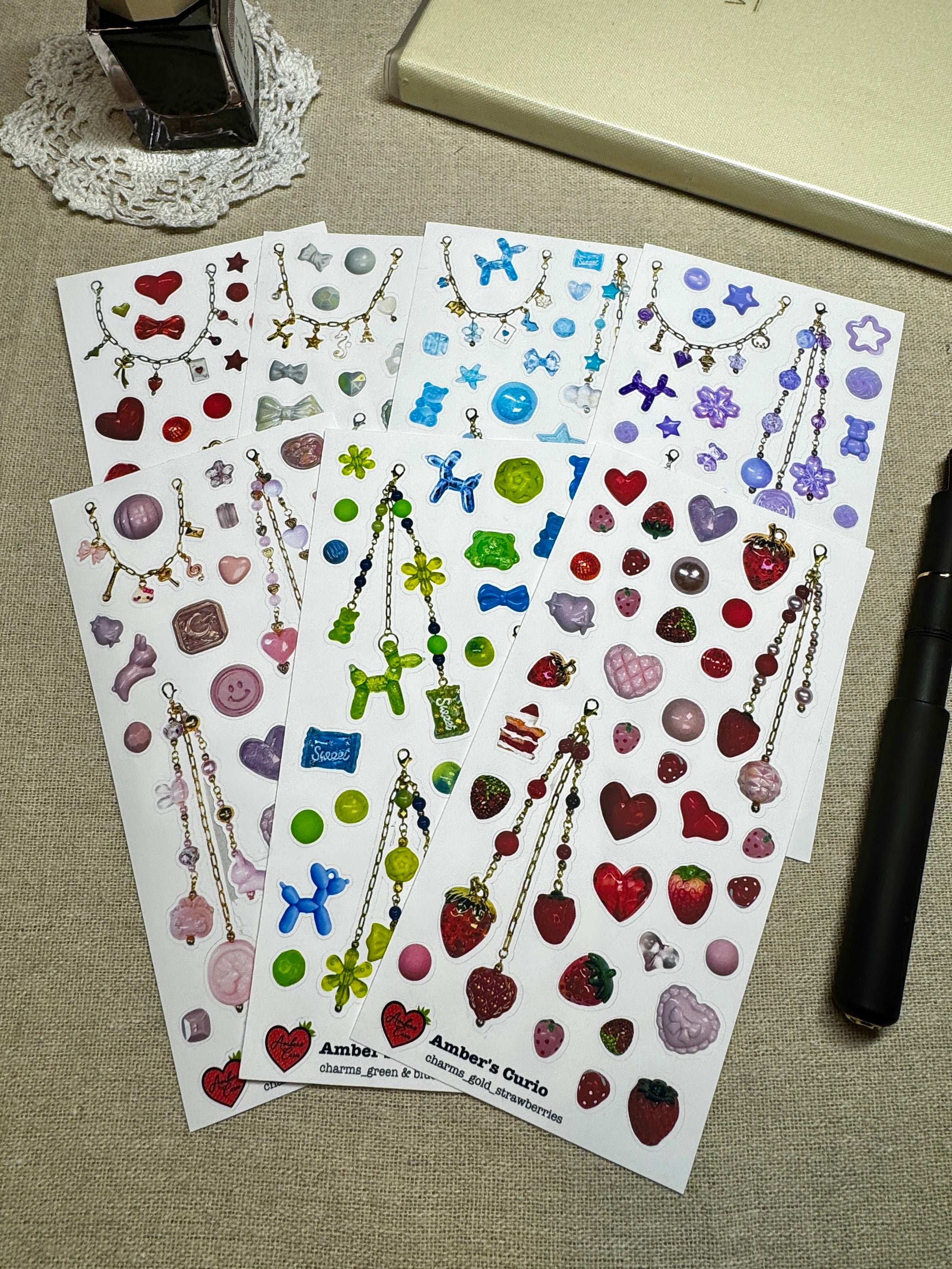 Sticker sheets of photographed beads and charms printed on premium matte paper styled on linen next to a black fountain pen and ink. 