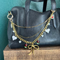 Gold chain with heart and bow charms draped over a black handbag on a teal surface.
