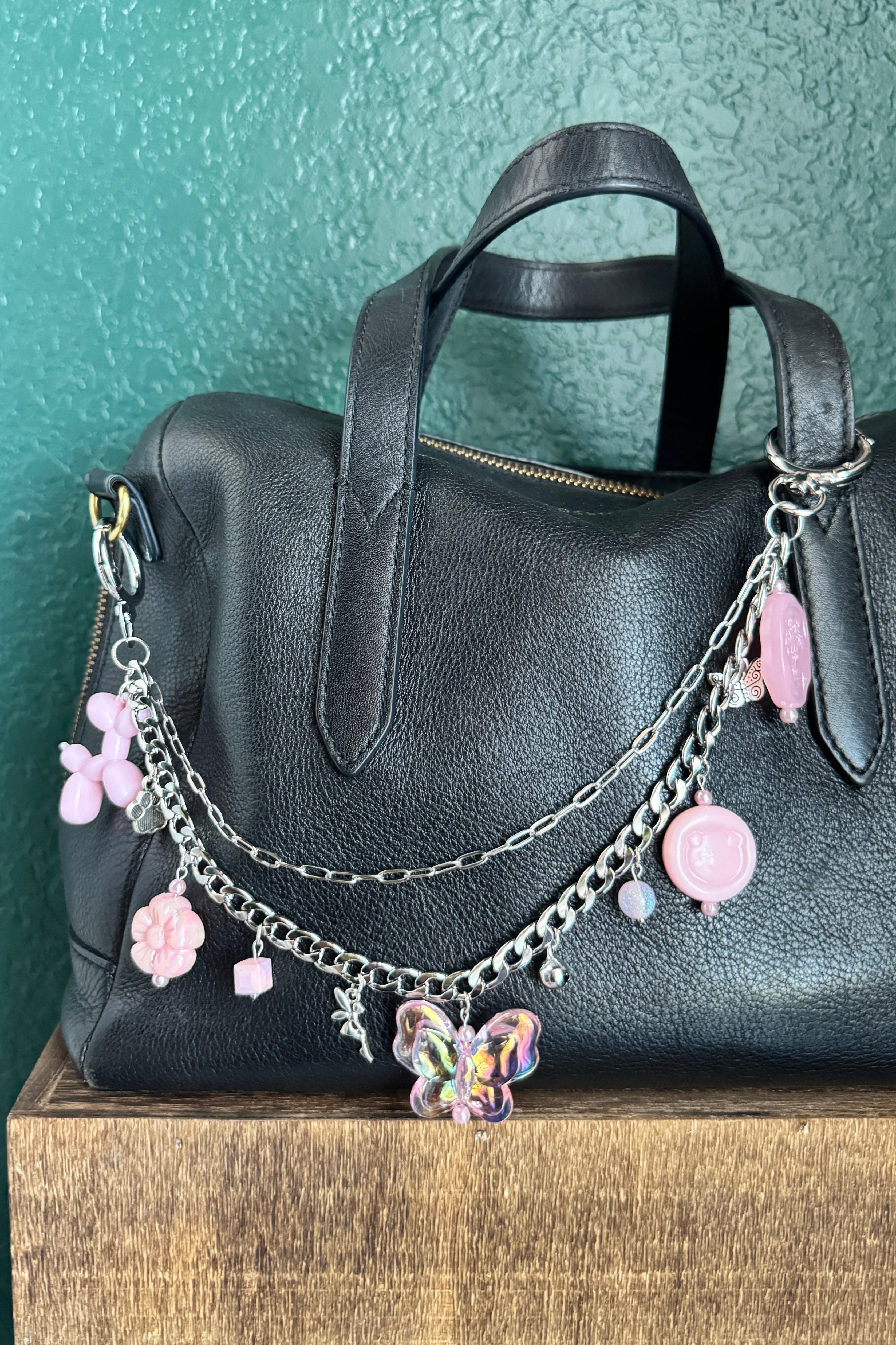 Black leather bag with decorative chain and pink charms on a textured green surface.