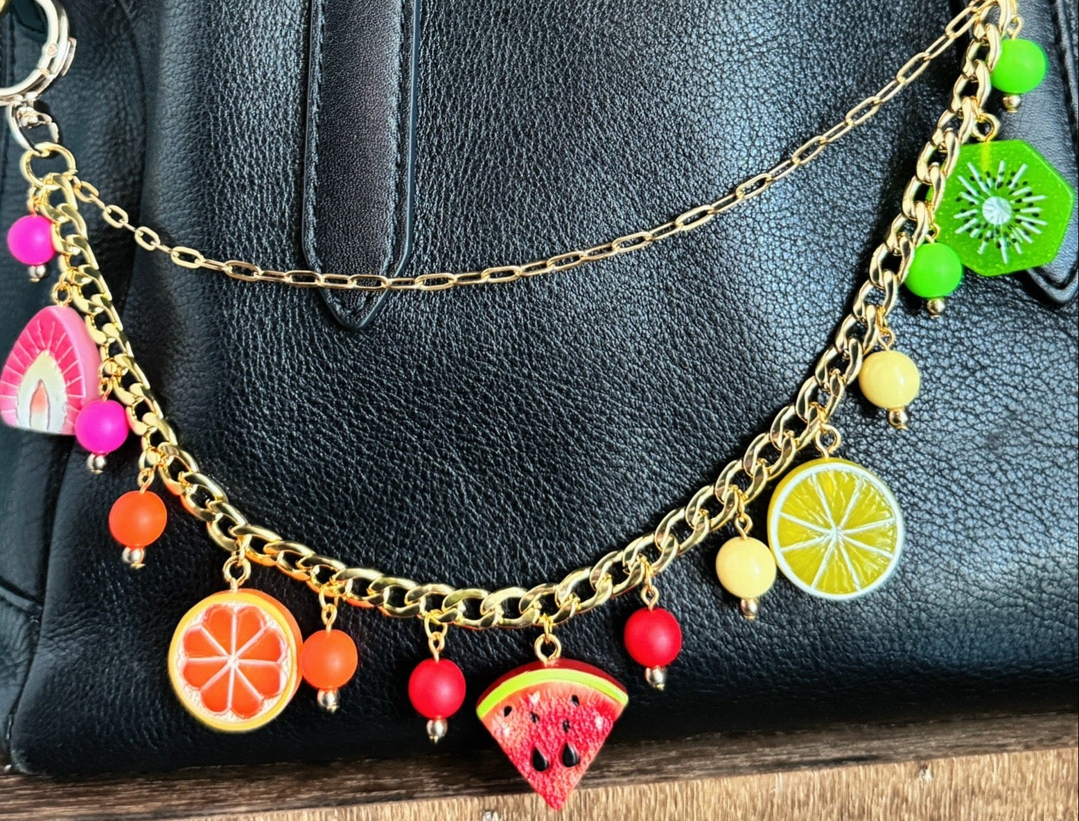 Black handbag with colorful fruit-themed charm chain against a teal wall.