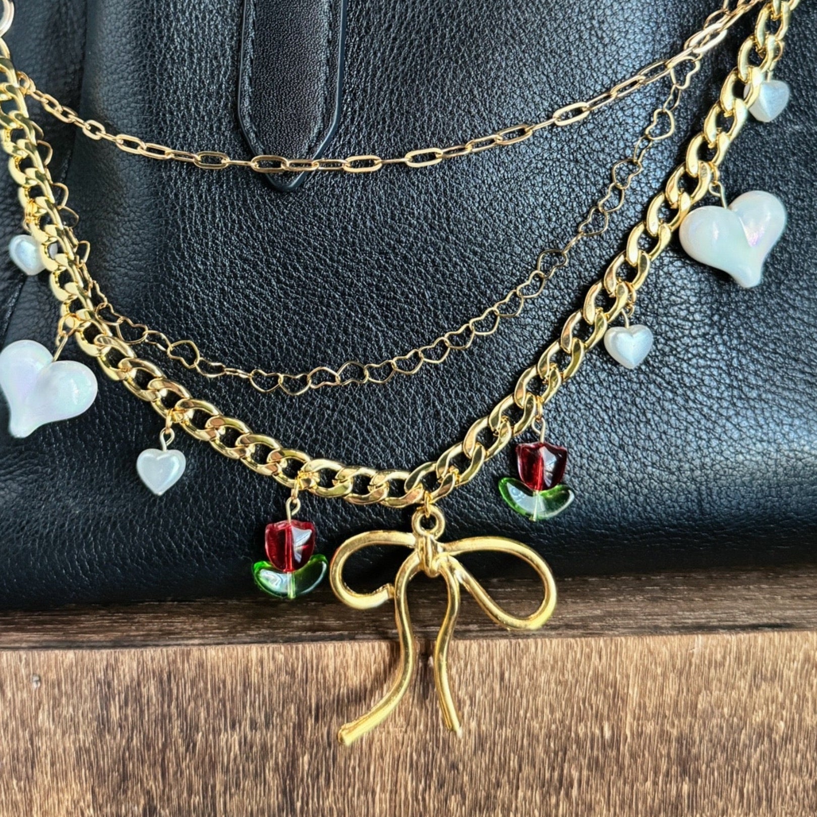 Gold chain necklace with heart and bow charms on a black handbag against a teal wall.