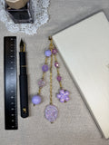 Purple decorative journal charm with beads and a gold chain on a gray surface with a pen and ruler.