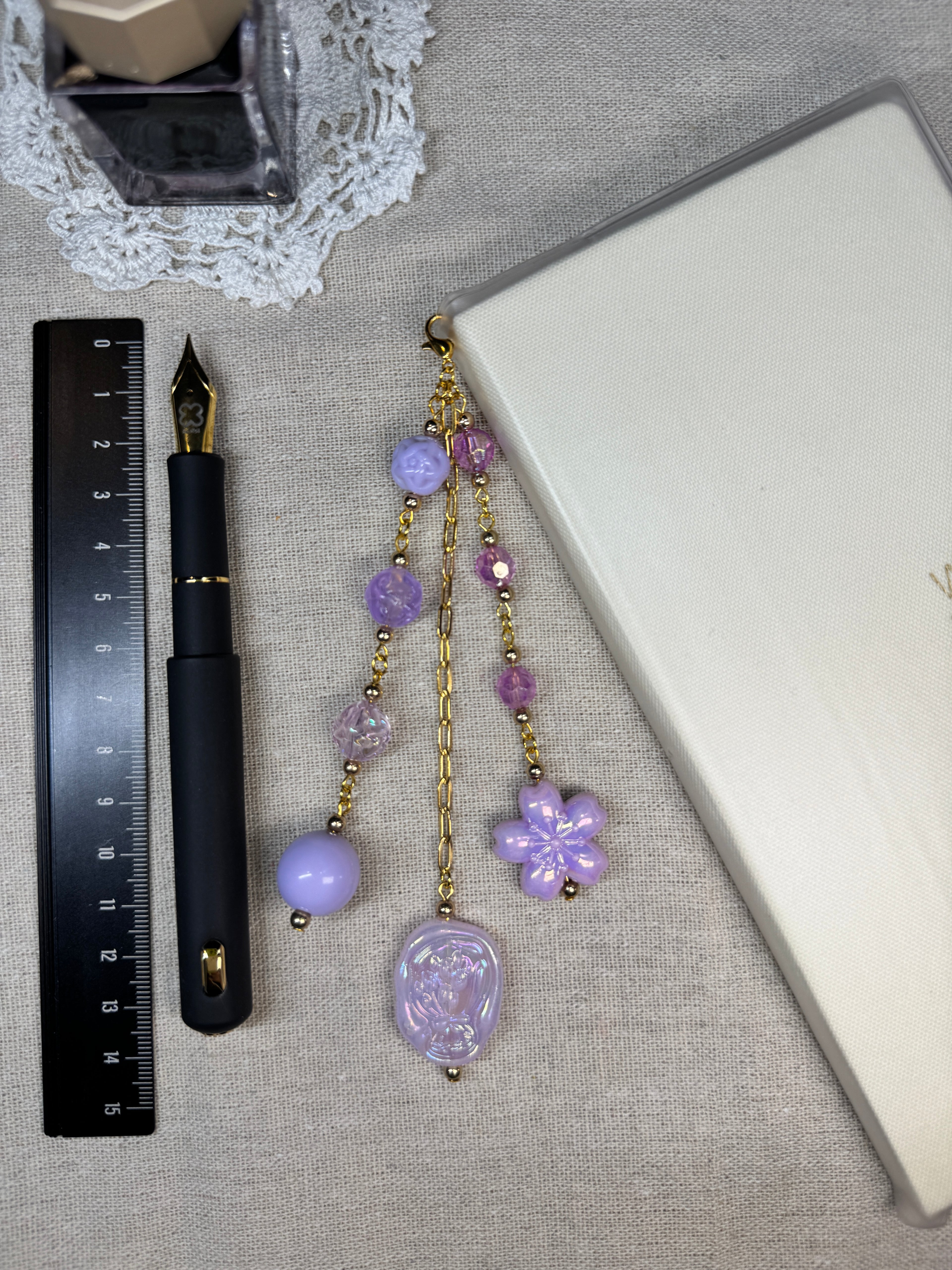 Purple decorative journal charm with beads and a gold chain on a gray surface with a pen and ruler.
