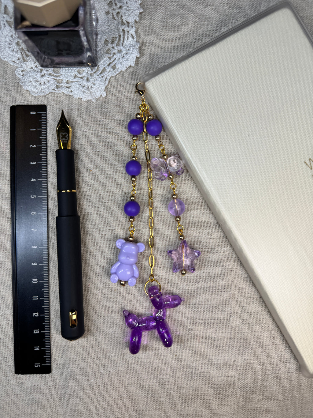 Decorative journal charm with purple and gold beads on a linen surface with a pen and ruler.