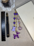 Decorative journal charm with purple and gold beads on a linen surface with a pen and ruler.