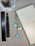 Decorative blue stars and clouds journal charm with a pen with charm on a textured surface next to a ruler and inkwell.
