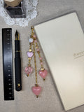Decorative light pink heart journal charm with pink and gold beads laid on top of linen surface with a pen and ruler.