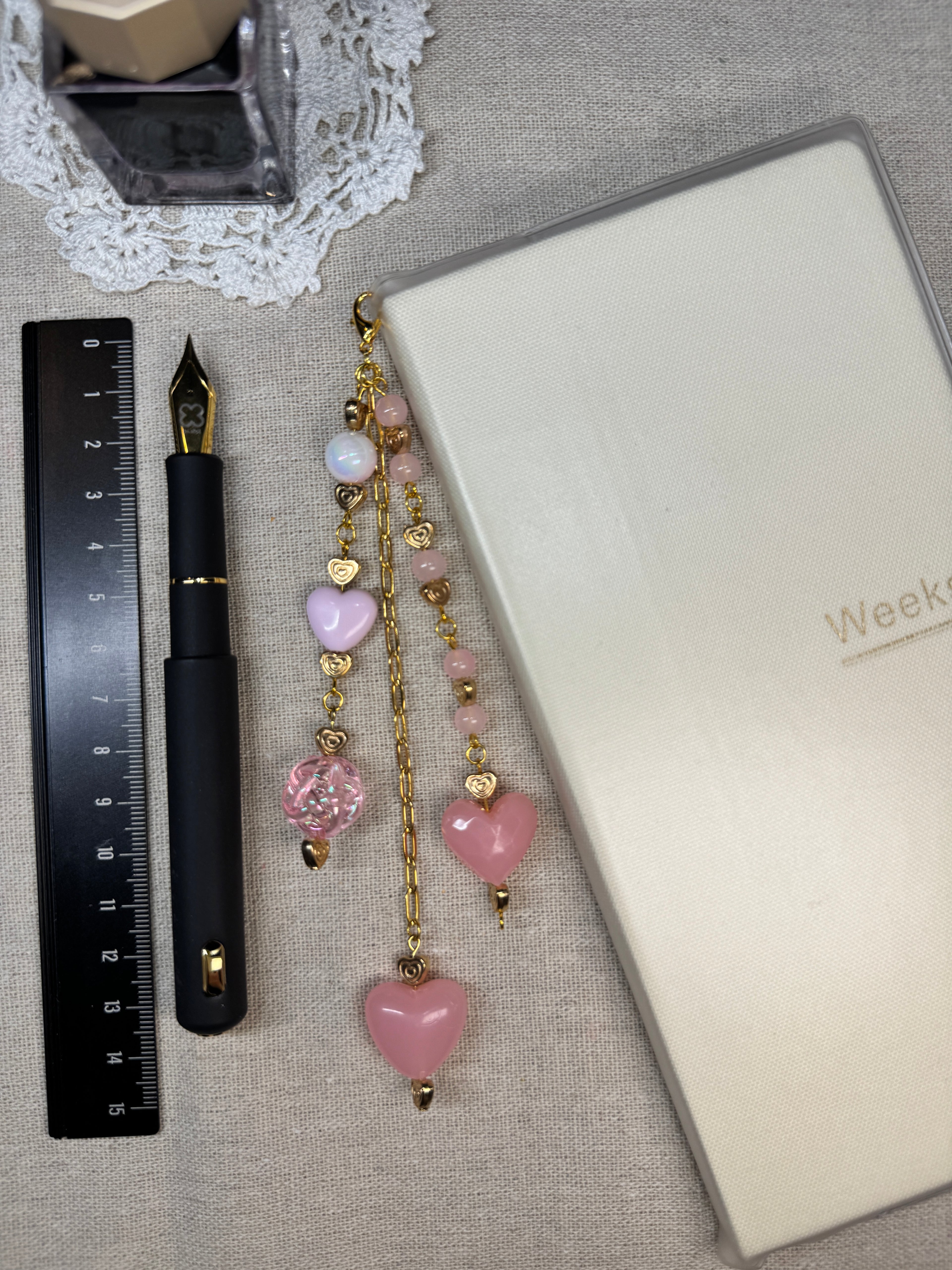 Decorative light pink heart journal charm with pink and gold beads laid on top of linen surface with a pen and ruler.