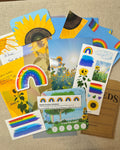 A flat lay of colorful sunflower and rainbow-themed cards, stickers, and a small kraft paper card on a beige fabric surface.