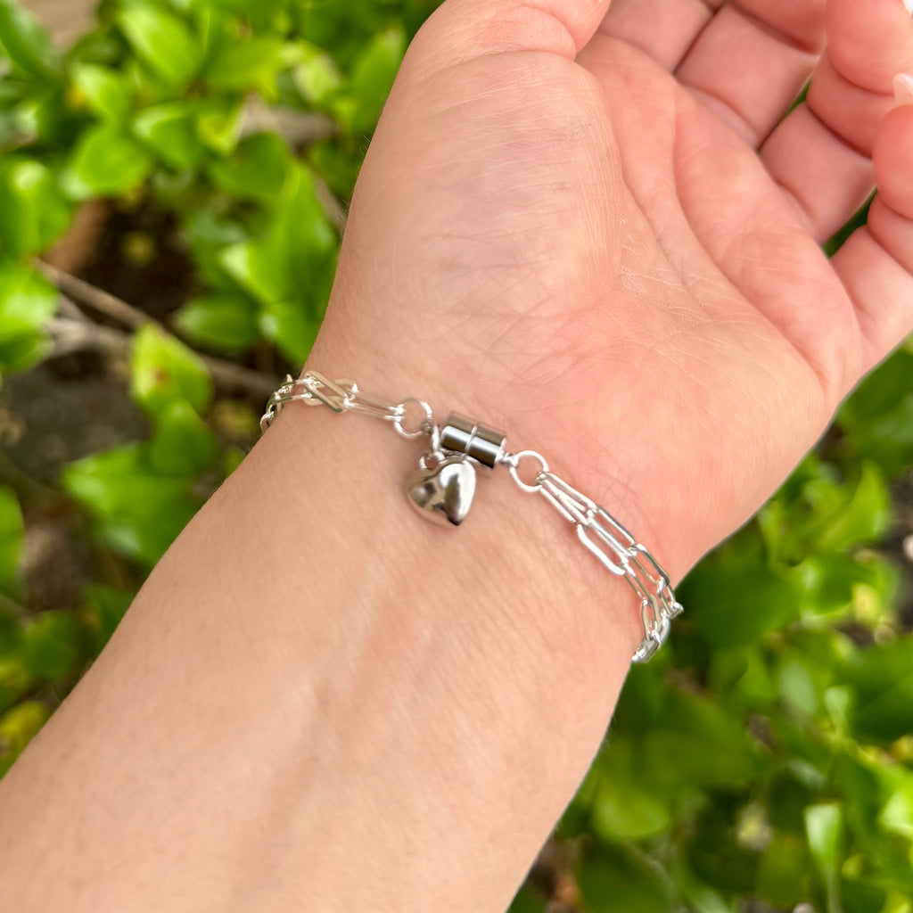 Silver bracelet with a heart charm on a person's wrist against a green leafy background