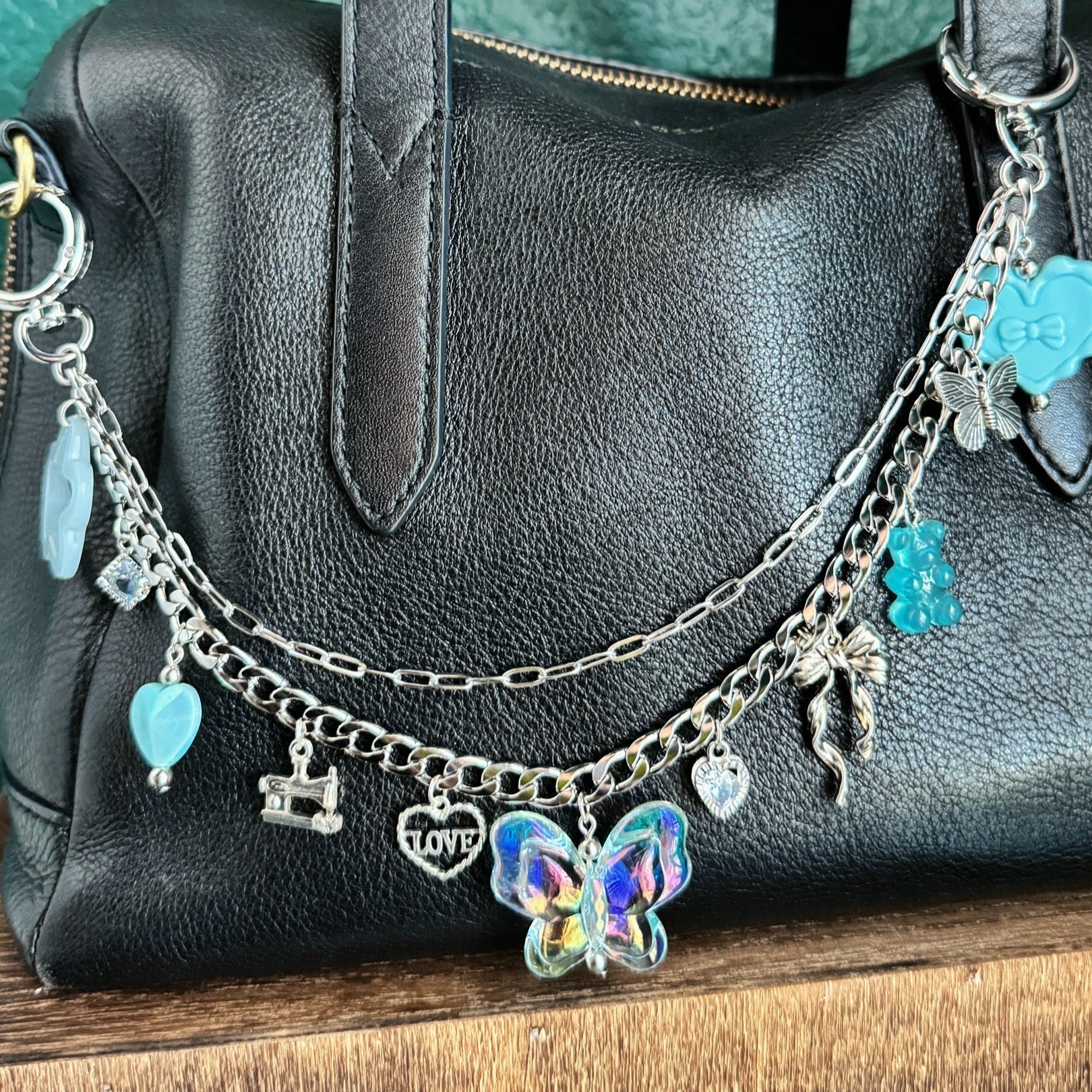 Black handbag with charm bracelets on a wooden surface against a teal wall.