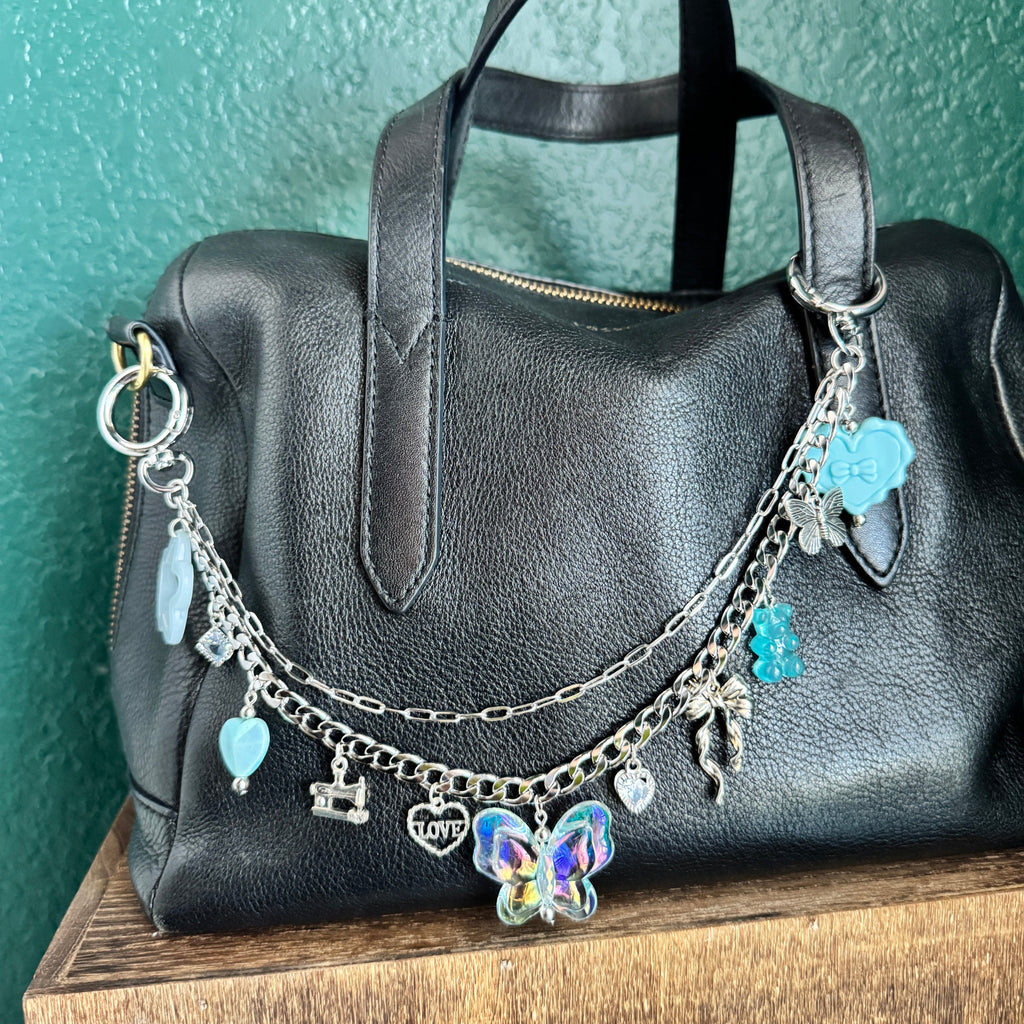Silver bag chain with blue acrylic charms on a black leather bag on top of a wooden box in front of a green wall 