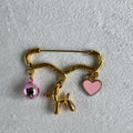 gold safety pin style brooch with a pink hear a balloon dog and a pink bell