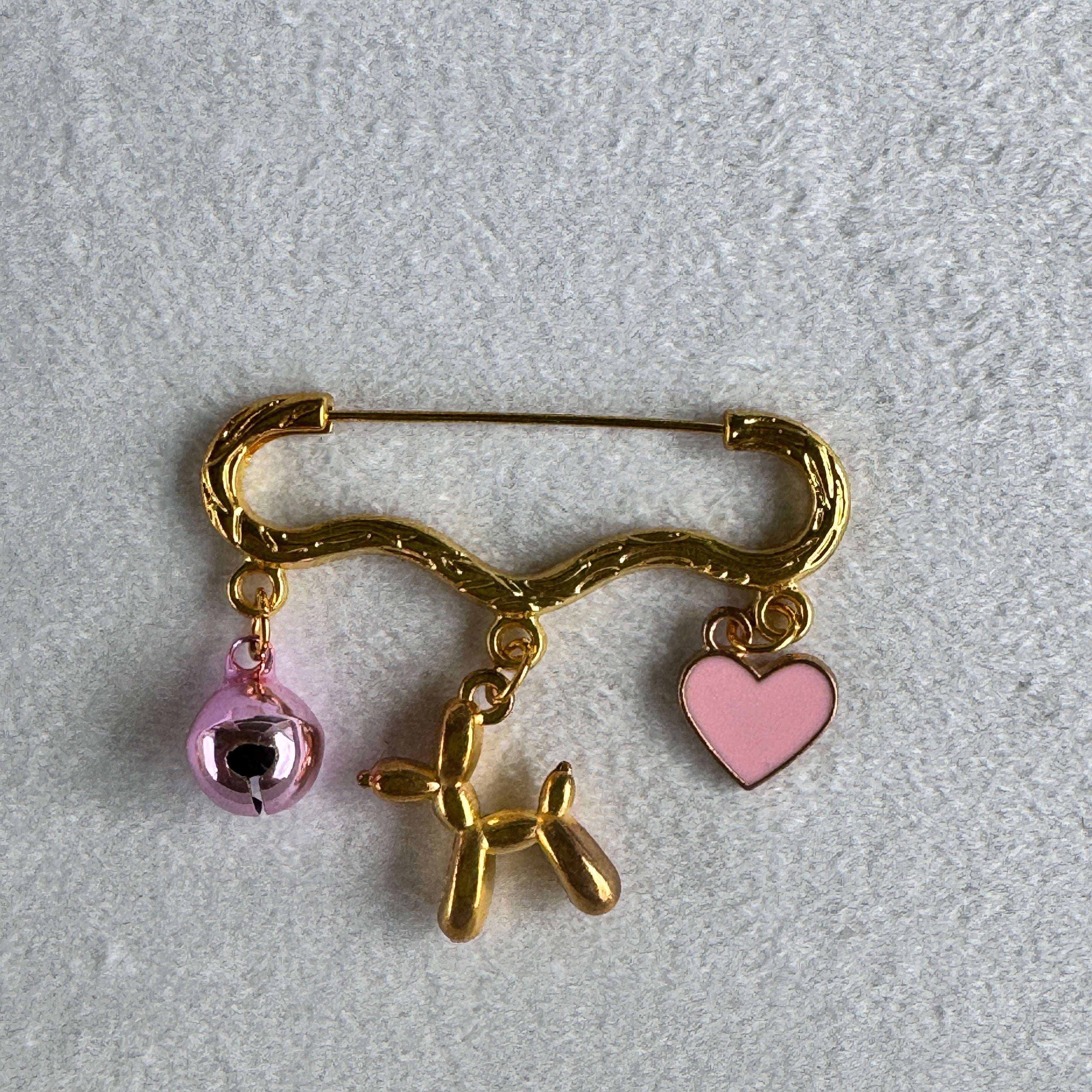gold safety pin style brooch with a pink hear a balloon dog and a pink bell